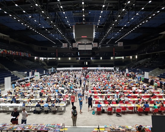 An indoor sports arena filled with rows of tables, each occupied by people engaged in competitive activities. The ceiling is high, with numerous lights illuminating the large space. The audience seating on either side is empty, and there are flags hanging from the upper level.